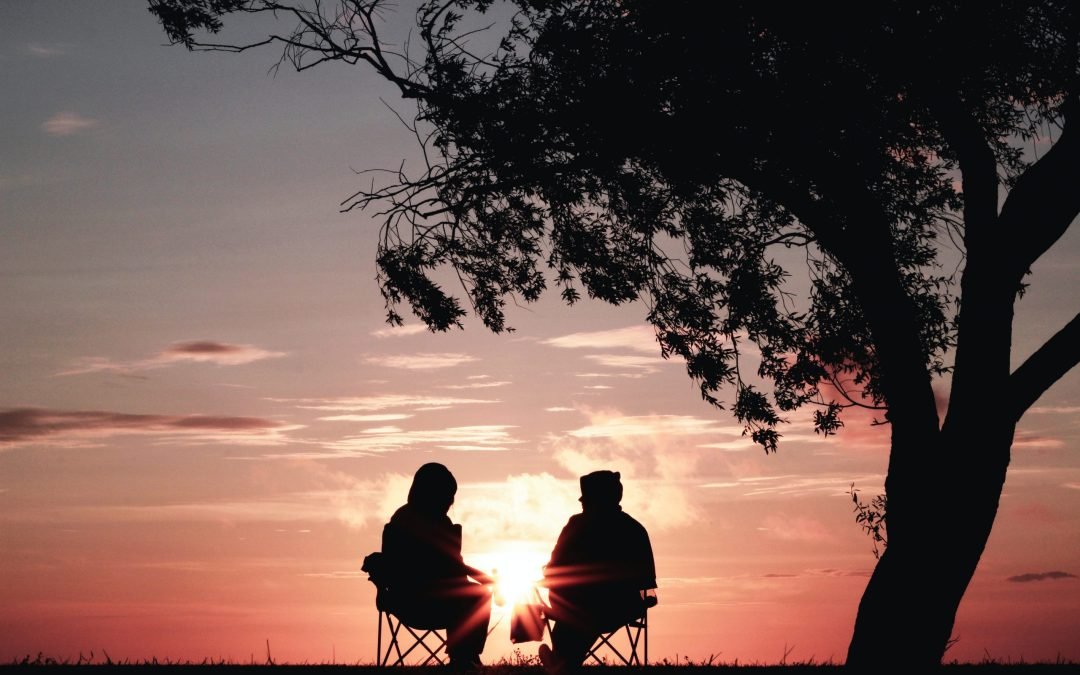 two silhouettes sitting in front of a tree at sunset